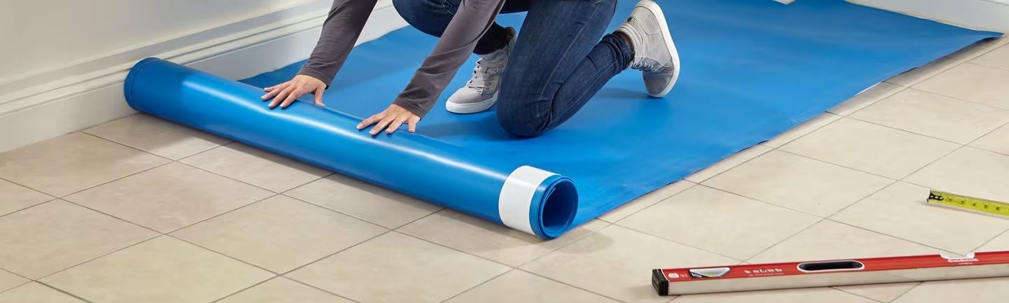 An installer kneeling to unroll blue underlayment across a tiled floor, a spirit level lying beside them.
