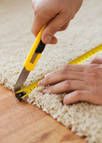 Hands drawing a trimming knife along a steel rule to cut cream carpet to the floor edge.