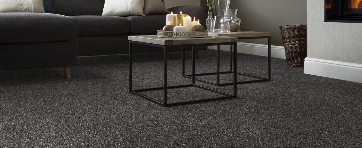 A lounge carpeted wall to wall in charcoal, a slim black coffee table set on it in front of a grey sofa.
