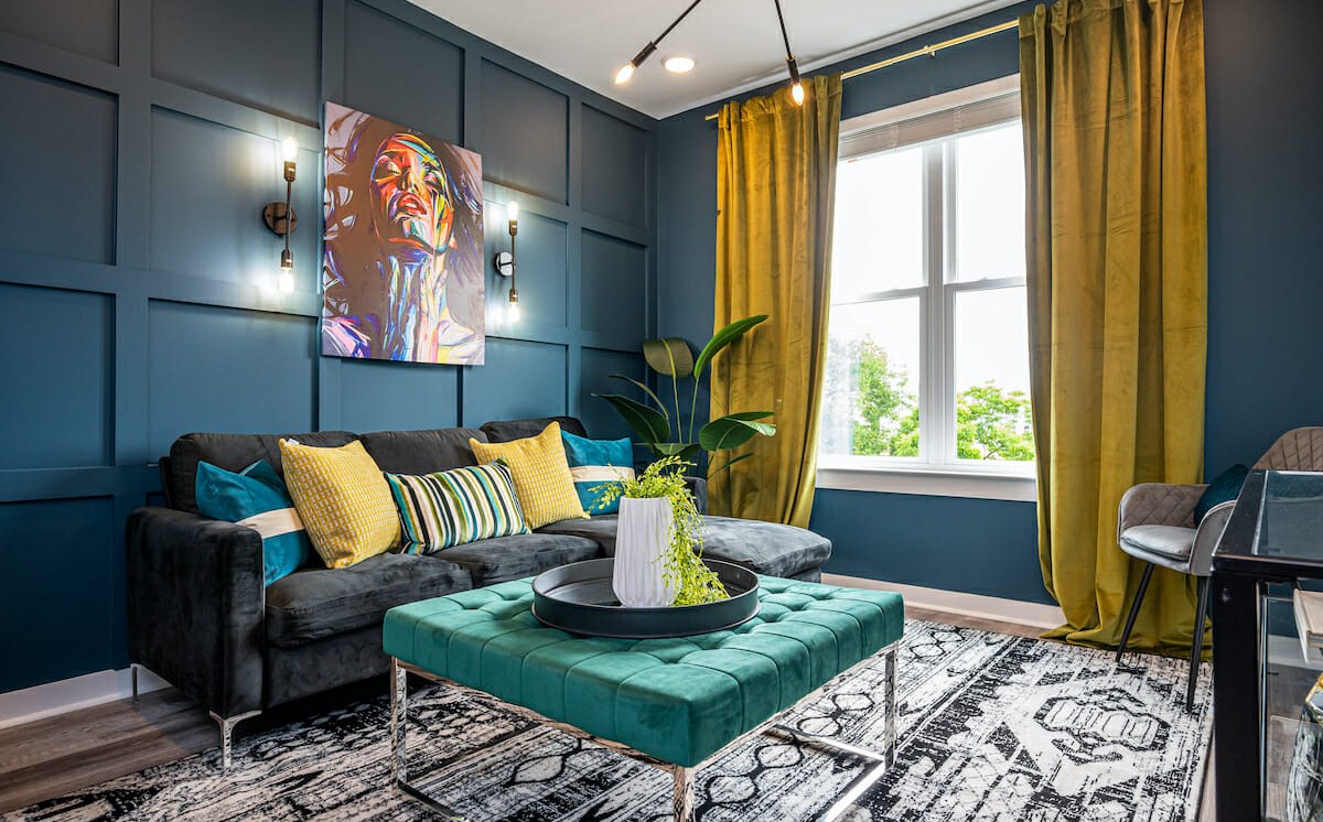 A finished blue-panelled lounge with a teal ottoman, yellow cushions and a patterned rug.