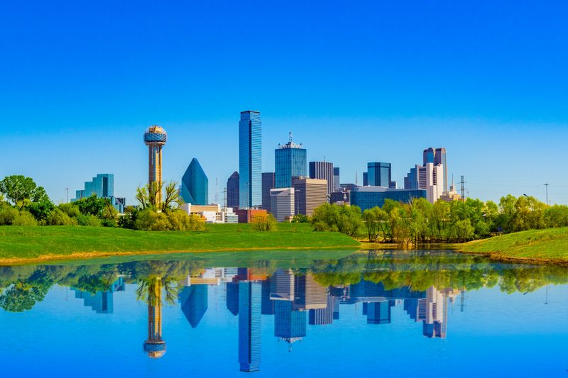 The Dallas skyline on a clear day, its towers mirrored in the still water of a lake in the foreground.