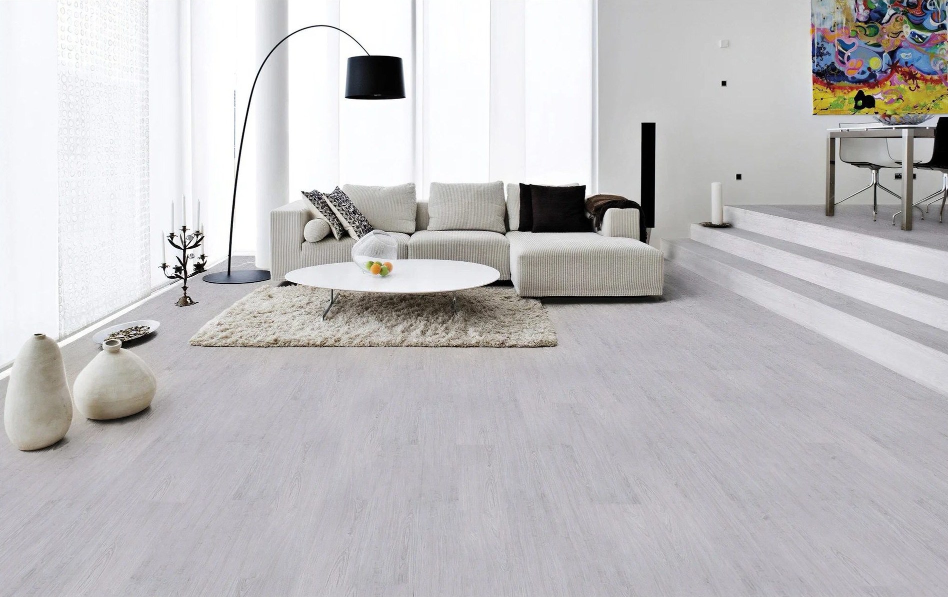 A bright, double-height living room with a pale corduroy sectional sofa and a round white coffee table, on wide light-grey plank flooring.