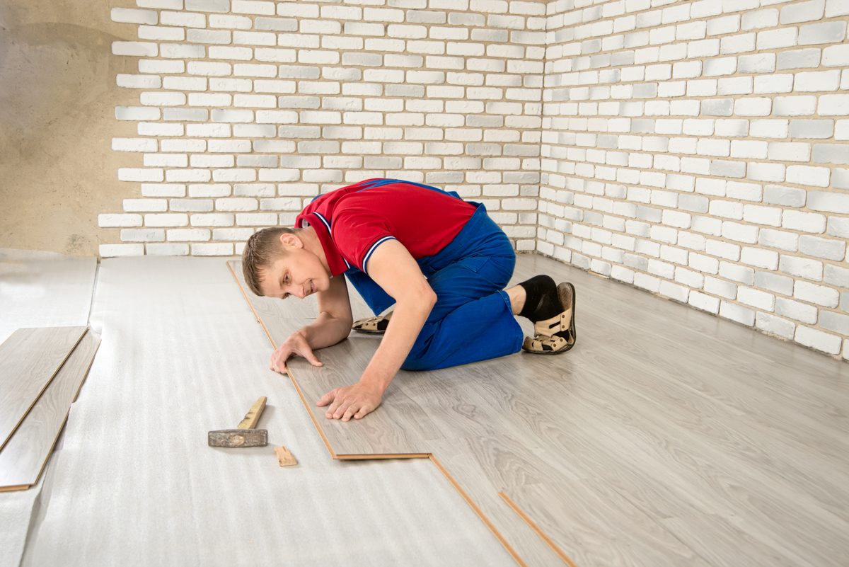 An installer kneeling on a new laminate floor, sliding a board into place against a white brick wall.