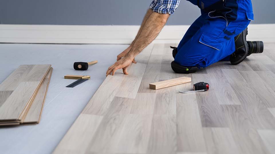 An installer in blue overalls kneeling to press a laminate board into place, a mallet, saw and tape measure beside him.