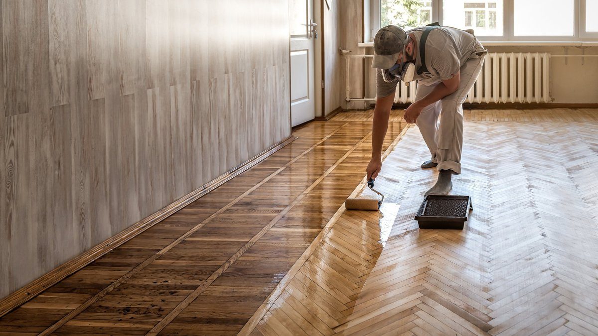 A contractor rolling varnish along a herringbone parquet floor in a bright hallway.
