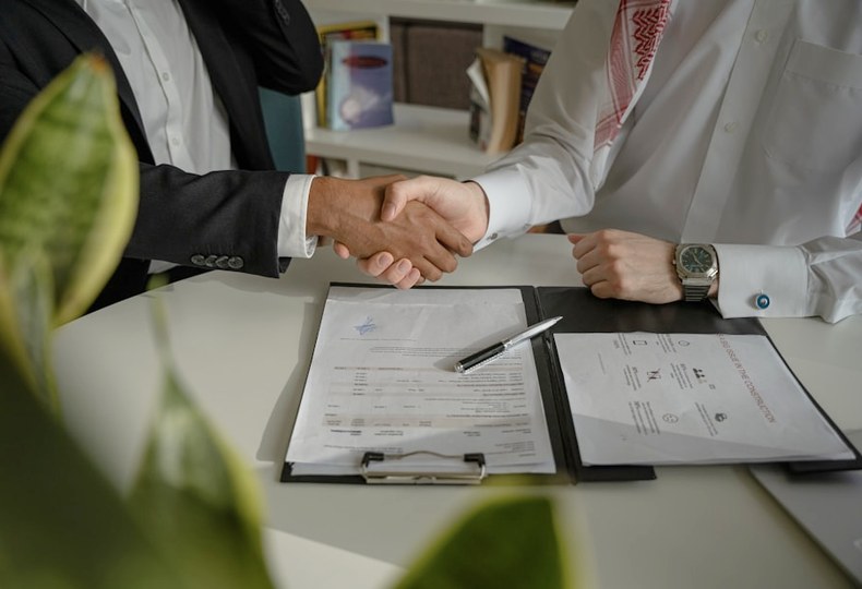 Two people shaking hands across a desk over a signed plan and a set of drawings.